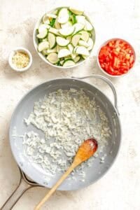 Process shot showing onions cooking in pan with veggies in separate bowls alongside pan.