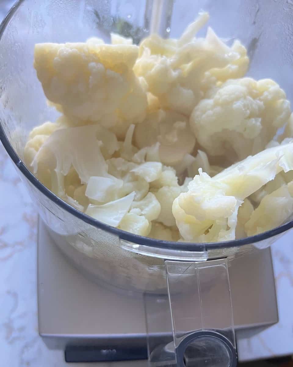 Cooked cauliflower florets piled in a food processor bowl, ready to blend into mashed cauliflower.