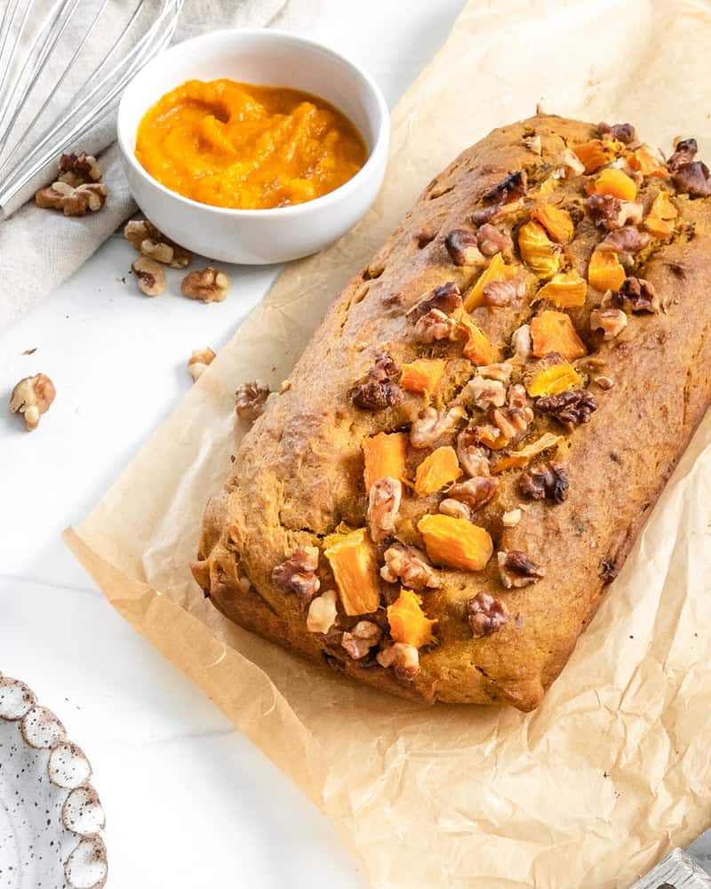 Whole vegan pumpkin bread loaf on parchment paper with a bowl of pumpkin puree.